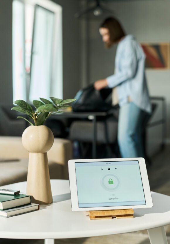 Caucasian young adult woman standing in modern living room packing bag in background, digital tablet with security lock screen displayed on table in foreground, plant and books nearby